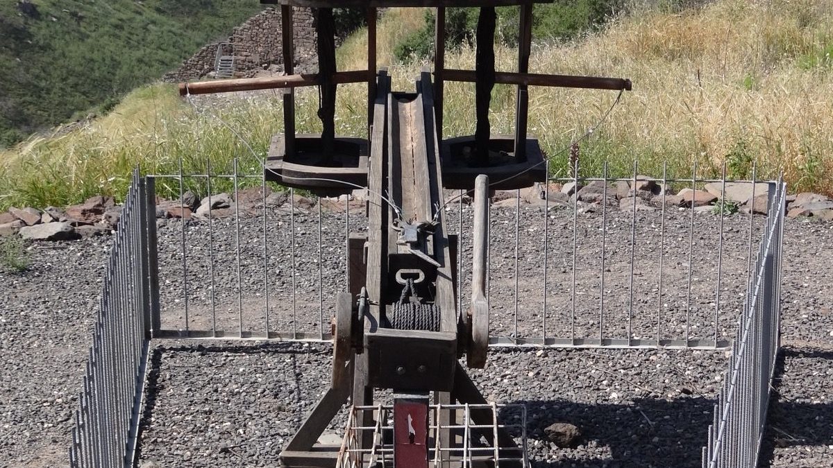Photograph of a reconstructed Roman ballista model at the Gamla Nature Reserve in Israel, showing its twin torsion arms and frame mounted above a stack of stone ammunition, with the Golan hillside in the background