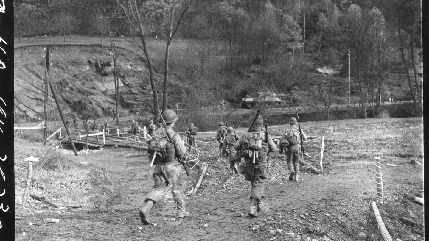 American infantrymen carrying rifles and bazookas cross a log footbridge into the Hürtgen Forest near a Sherman tank, November 18, 1944