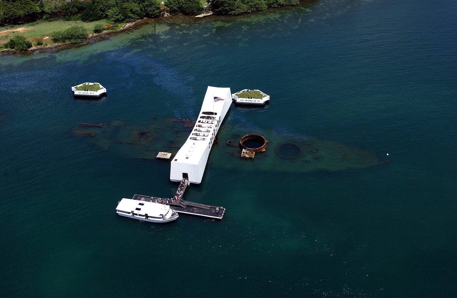 Aerial view of the white USS Arizona Memorial spanning the sunken hull at Pearl Harbor, with a Navy tour boat moored at its pier